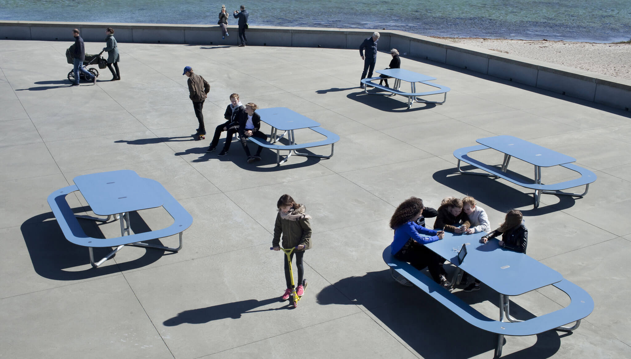 Fem blå PLATEAU Picnic O bordbænkesæt placeret på stranden med mennesker, der nyder siddepladserne