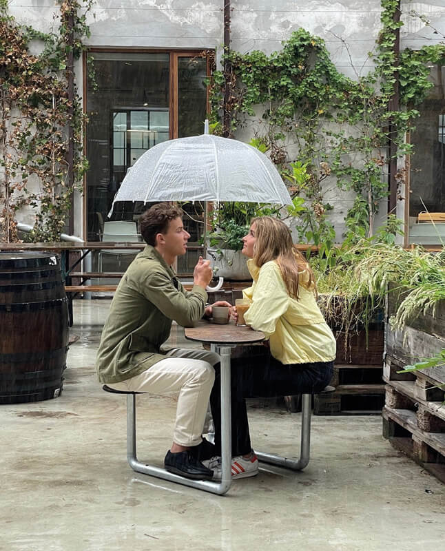 To personer der sidder i læ under paraplyen på et Plateau Picnic Duo bordbænkesæt på en cafe