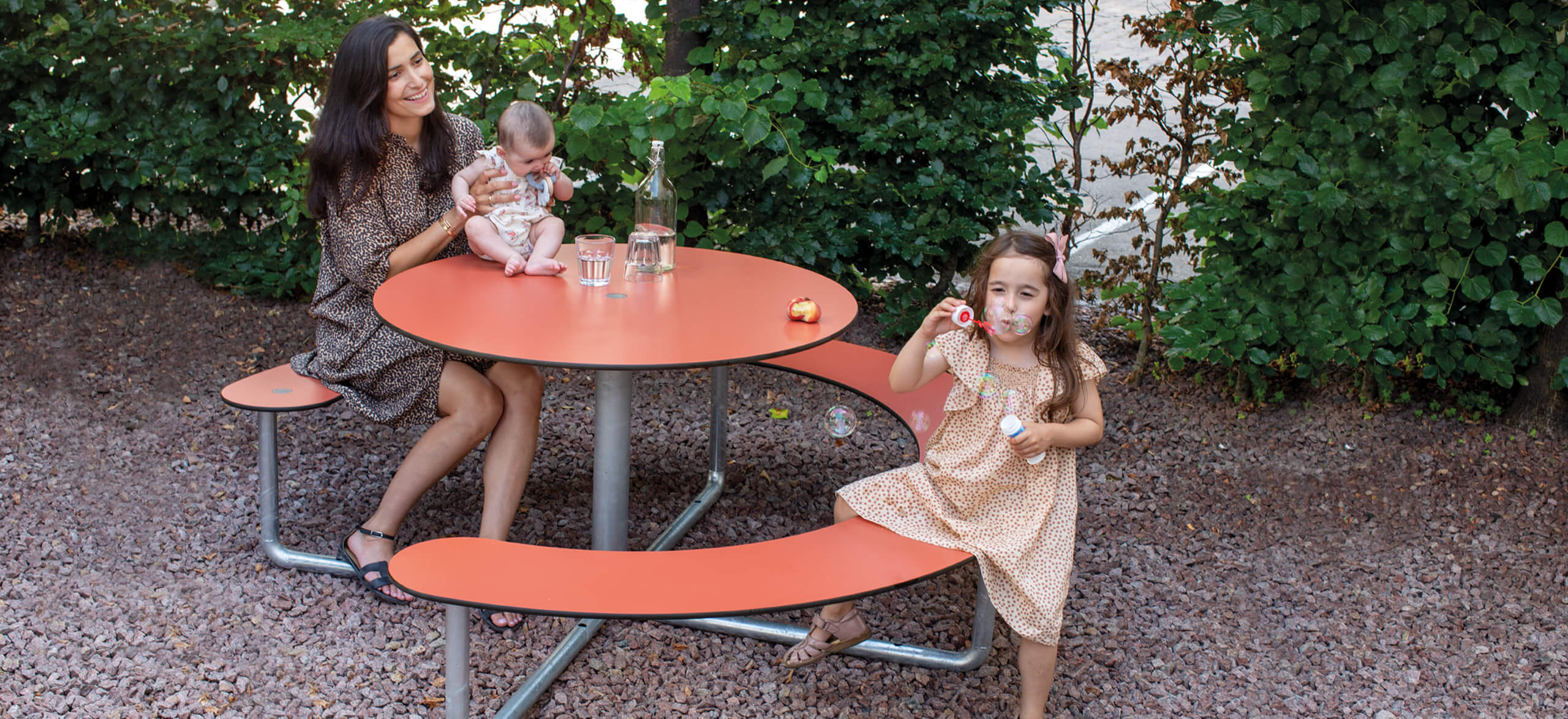 En mor og hendes to børn ved et Plateau Picnic Round bordbænkesæt i en park. Den ene af børnene puster sæbebobler.
