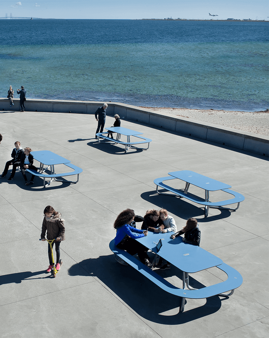 Tre blå PLATEAU Picnic O bordbænkesæt ved stranden, hvor 6 personer tager en velfortjent siddepause